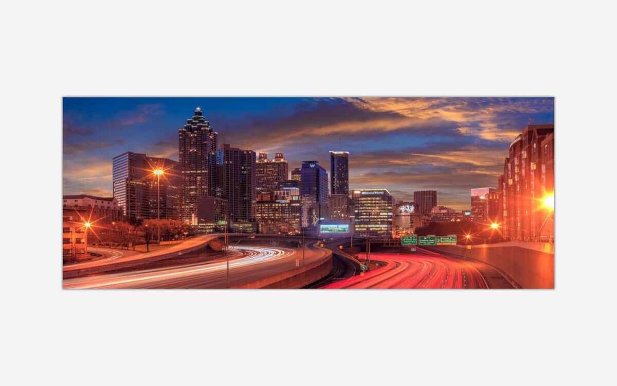 A panoramic view of a city skyline at dusk with illuminated buildings and light trails from moving vehicles on the highway.