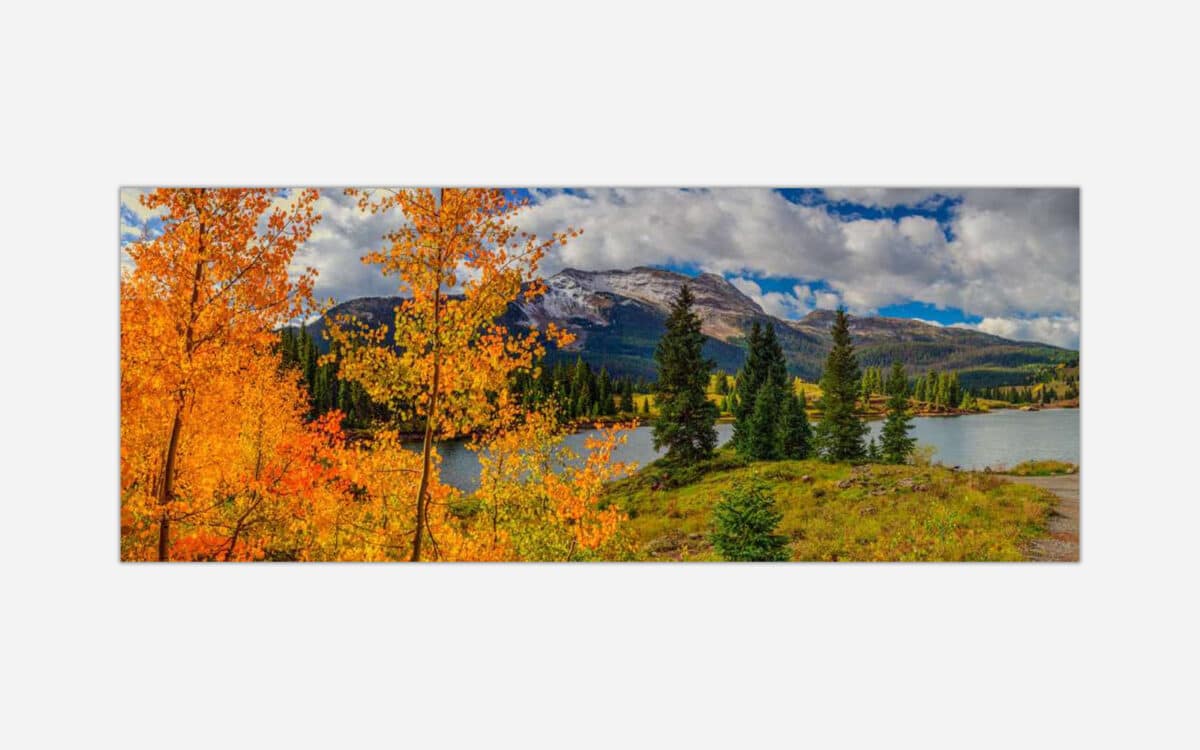 A panoramic autumn landscape with vibrant orange and yellow foliage, a serene lake, pine trees, and a mountain background under a cloudy sky.