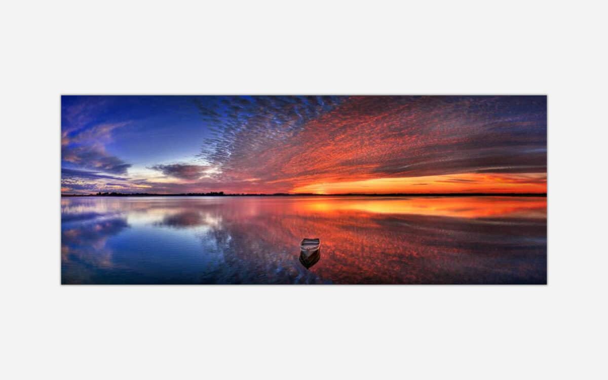 A panoramic painting of a tranquil lake sunset with vibrant sky colors reflected in the water and a lone boat.
