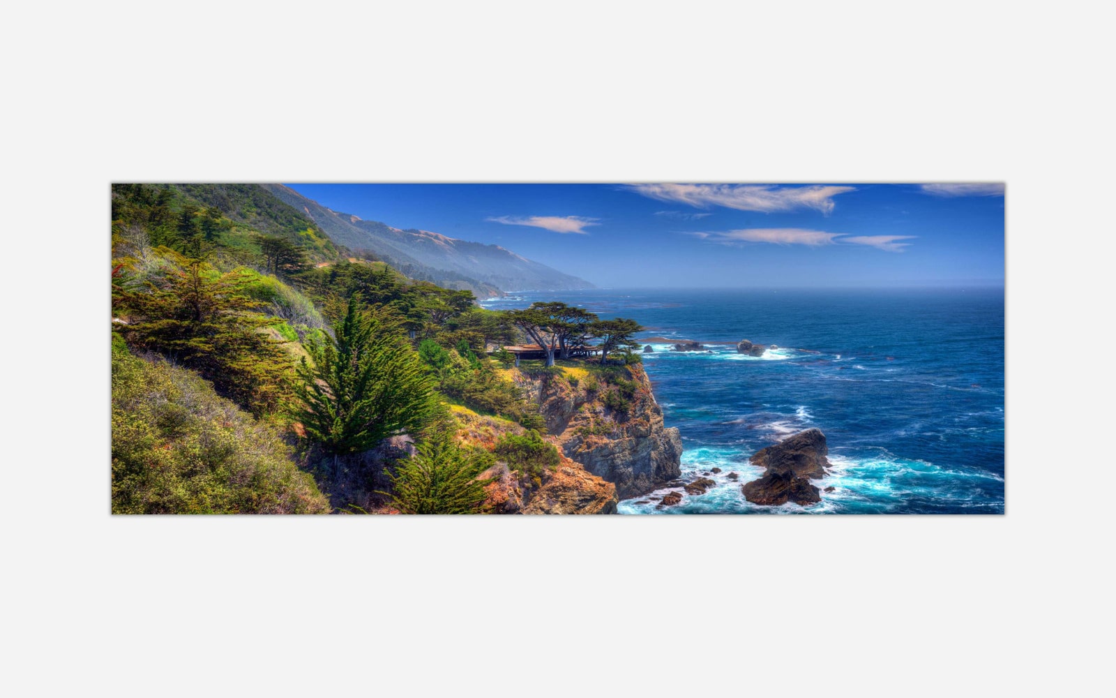 Big Sur (1) A panoramic view of a coastal landscape with a rocky shoreline, blue ocean, greenery, and a clear sky.