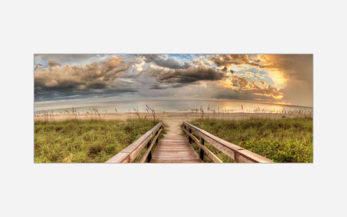 A panoramic art print of a serene beach scene with a wooden path leading to the ocean, featuring lush beach grass, a cloudy sky, and a warm sunset glow.