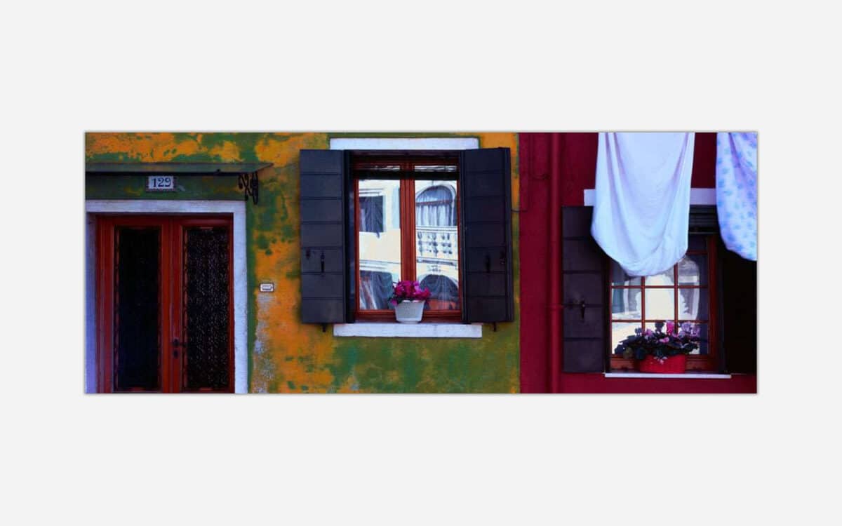 A photograph capturing a colorful urban scene with a green and orange textured wall, a red door, and two windows, one adorned with flowers and the other with hanging laundry.