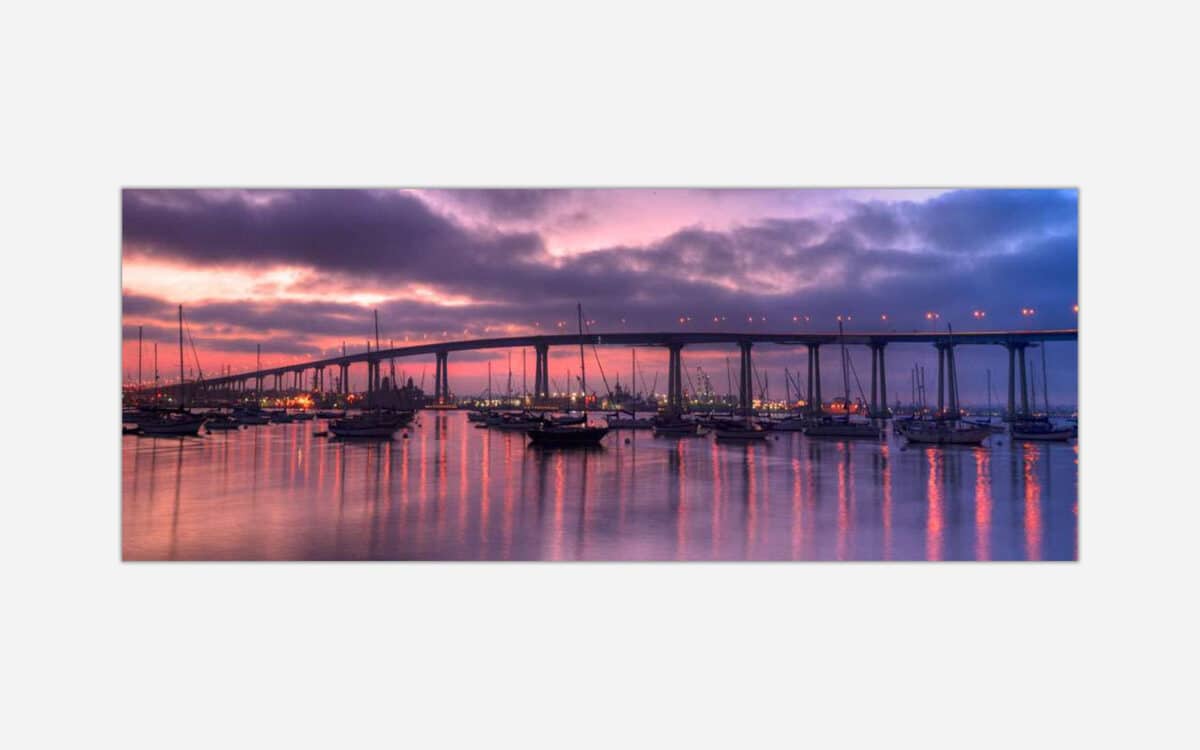 A panoramic photograph of a marina with boats and a bridge in the background under a purple sunset sky.