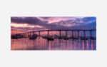 A panoramic photograph of a marina with boats and a bridge in the background under a purple sunset sky.