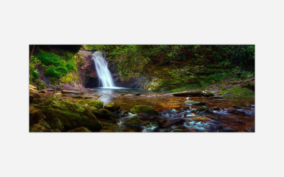 A panoramic photograph of a small serene waterfall in a lush forest setting with vibrant greens and flowing water over river rocks.