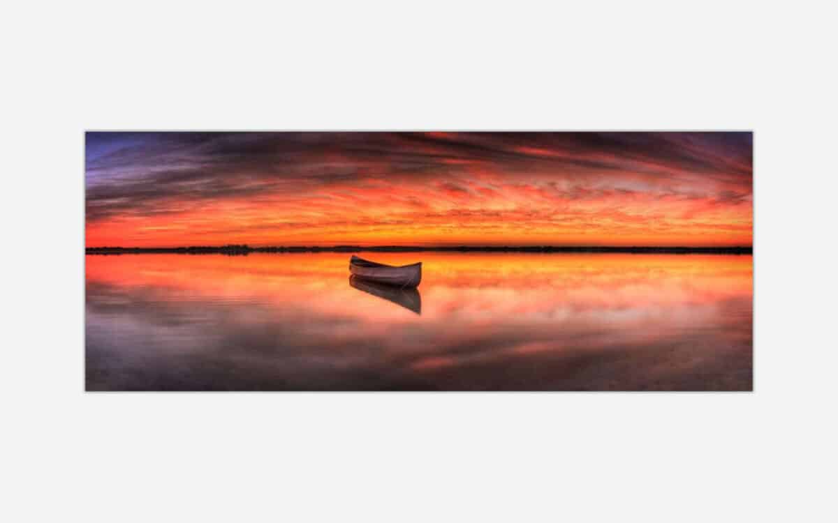 A photographic art piece featuring a serene lake with a solitary boat, vibrant sunset skies with clouds reflected in the calm waters.