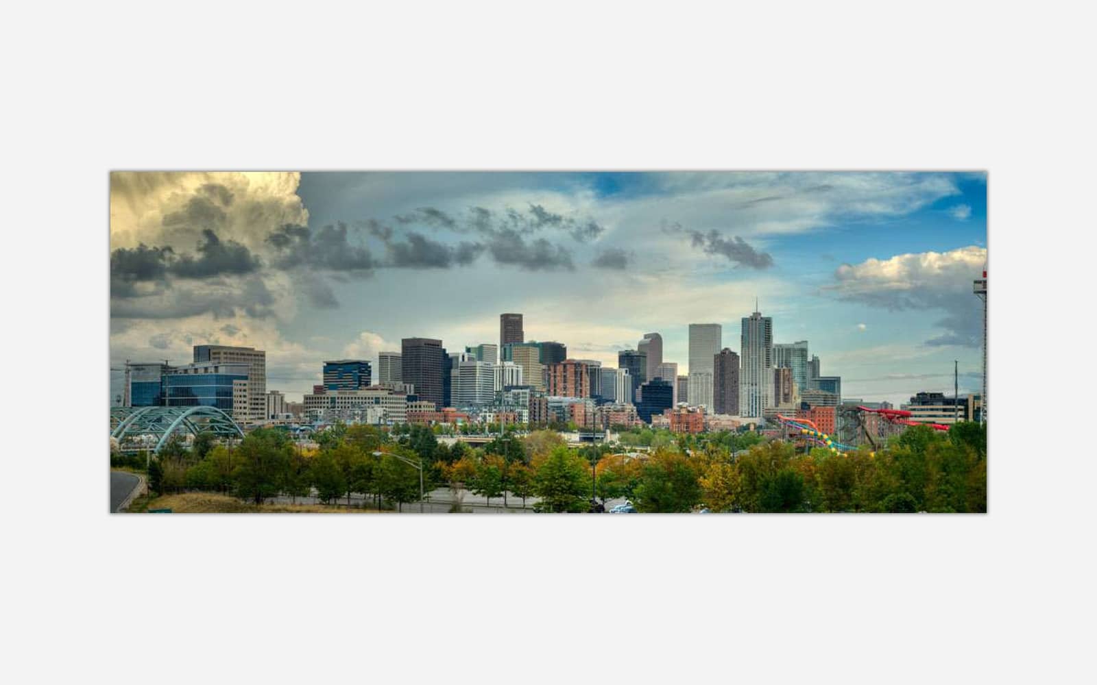 Denver A panoramic photograph of a modern city skyline with skyscrapers, dynamic clouds overhead, and greenery in the foreground.
