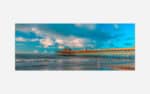 A vibrant photograph of a wooden pier extending into the ocean with waves lapping the shore under a blue sky with scattered clouds.