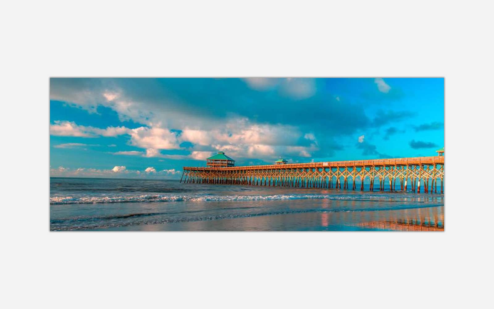 Folly Beach Pier (1) A vibrant photograph of a wooden pier extending into the ocean with waves lapping the shore under a blue sky with scattered clouds.