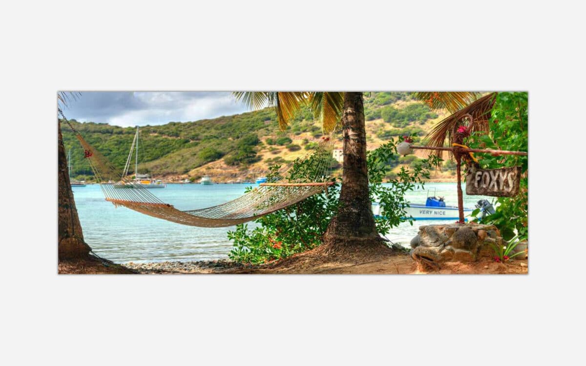 A picturesque view of a beach hammock tied to a palm tree with a sailboat in the distance, a sign for "Foxy's" bar, and the calm blue sea in the background.