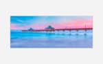 A panoramic photograph of a serene seascape with a pier extending into calm blue waters under a pastel pink and blue sunset sky.