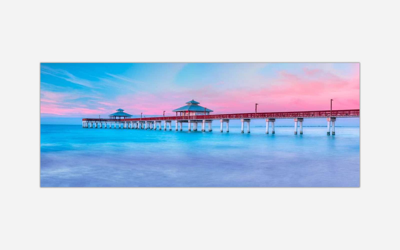 Ft. Myers II (1) A panoramic photograph of a serene seascape with a pier extending into calm blue waters under a pastel pink and blue sunset sky.