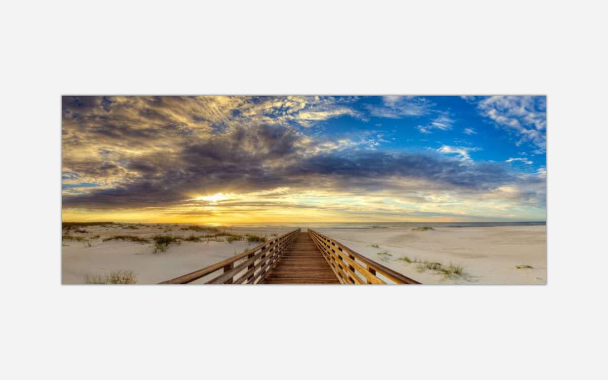 A panoramic image of a wooden walkway leading to a sunset on the beach with vibrant clouds and dune grass.