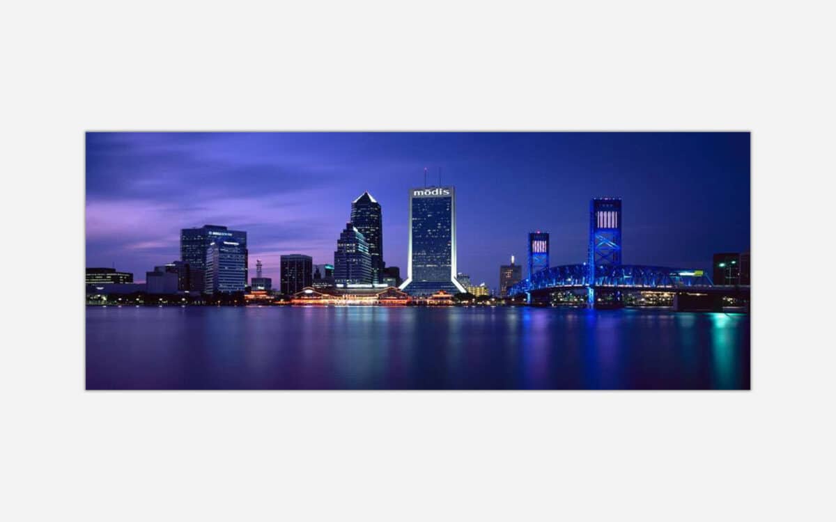 A panoramic view of a city skyline at twilight with illuminated high-rise buildings and a distinctive blue bridge reflected on the water's surface.