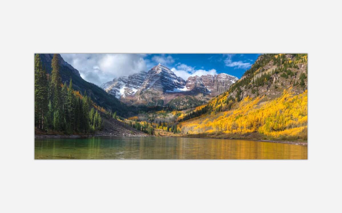 A panoramic photograph of a mountainous landscape with golden yellow aspen trees in autumn and a calm alpine lake in the foreground.