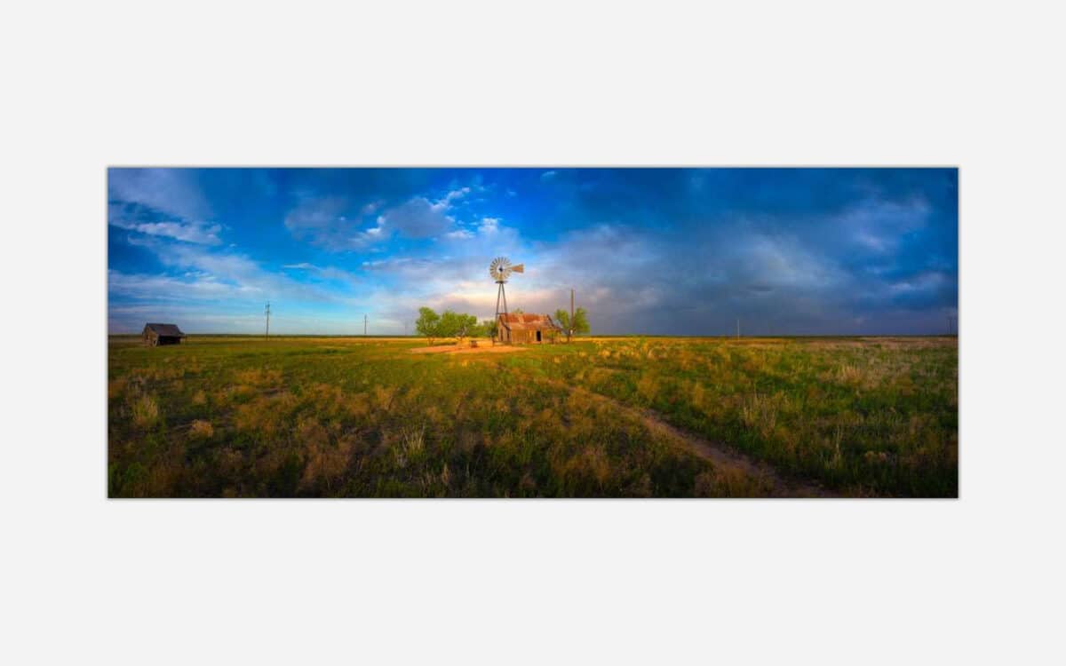 A photograph of an open rural landscape featuring a traditional windmill and old barn under a vast sky with warm sunset light.