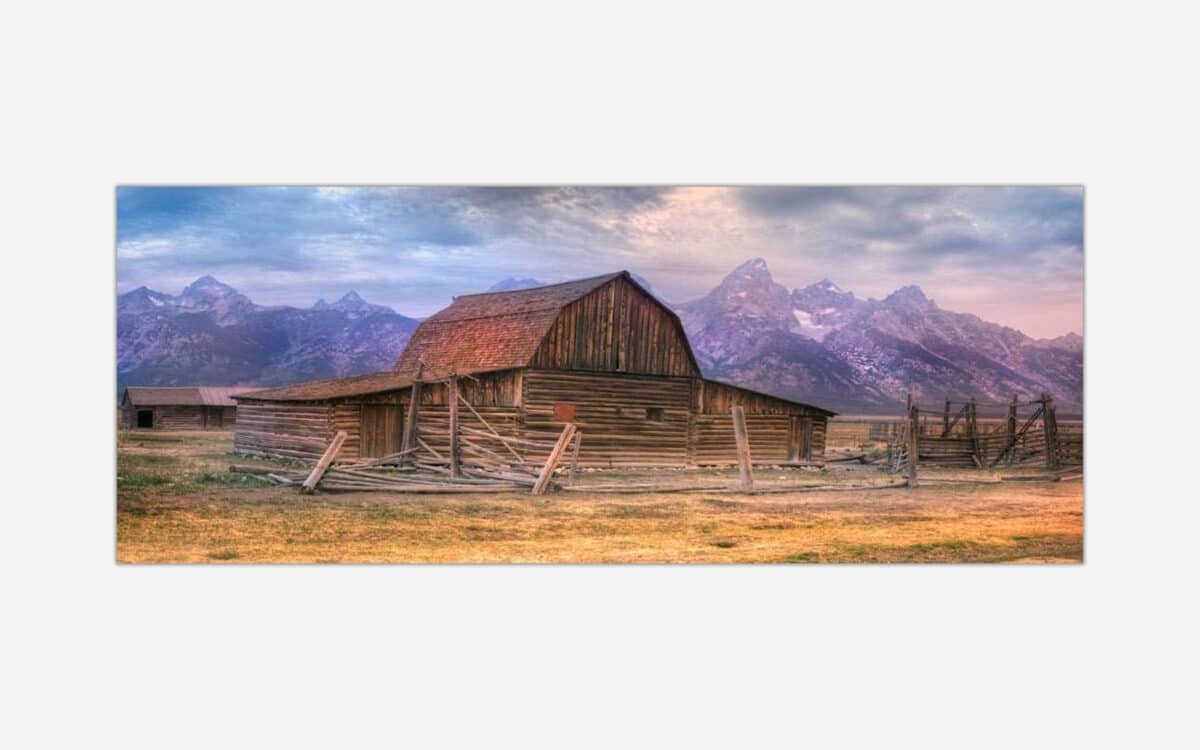 A rustic barn with a wooden fence set against a backdrop of majestic mountains and a warm, colorful sky.