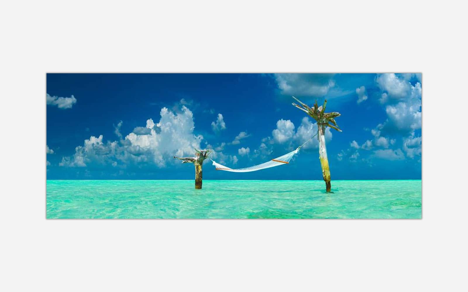 Paradise Found (1) A panoramic wall art of a hammock tied between two palm trees over crystal clear turquoise water under a blue sky with fluffy clouds.