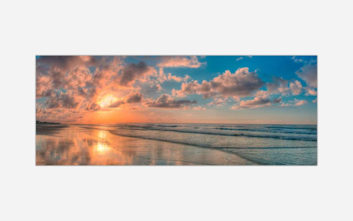 A scenic view of a beach at sunset with a vibrant sky reflected on the ocean and wet sand.