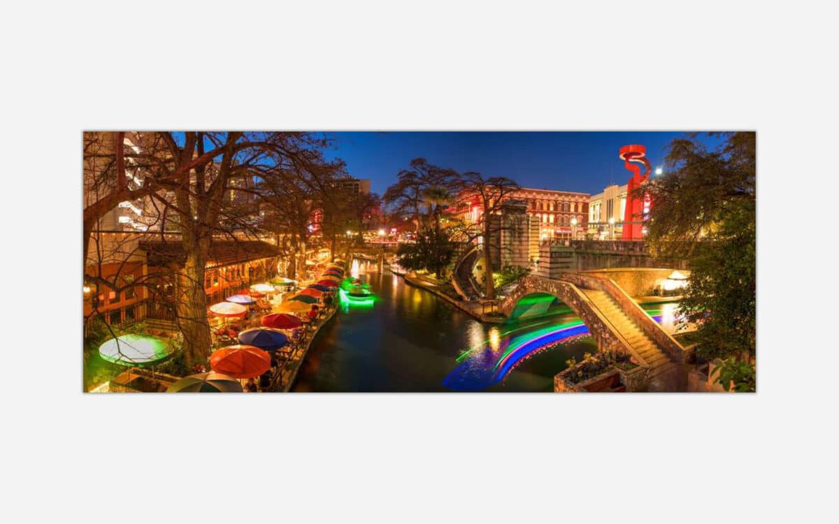 A vibrant night photograph of a city riverwalk area with illuminated bridges, colorful umbrellas, and lively outdoor restaurants.