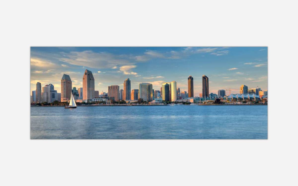 A panoramic photograph of a modern city skyline with skyscrapers and a sailboat on the water at sunset.