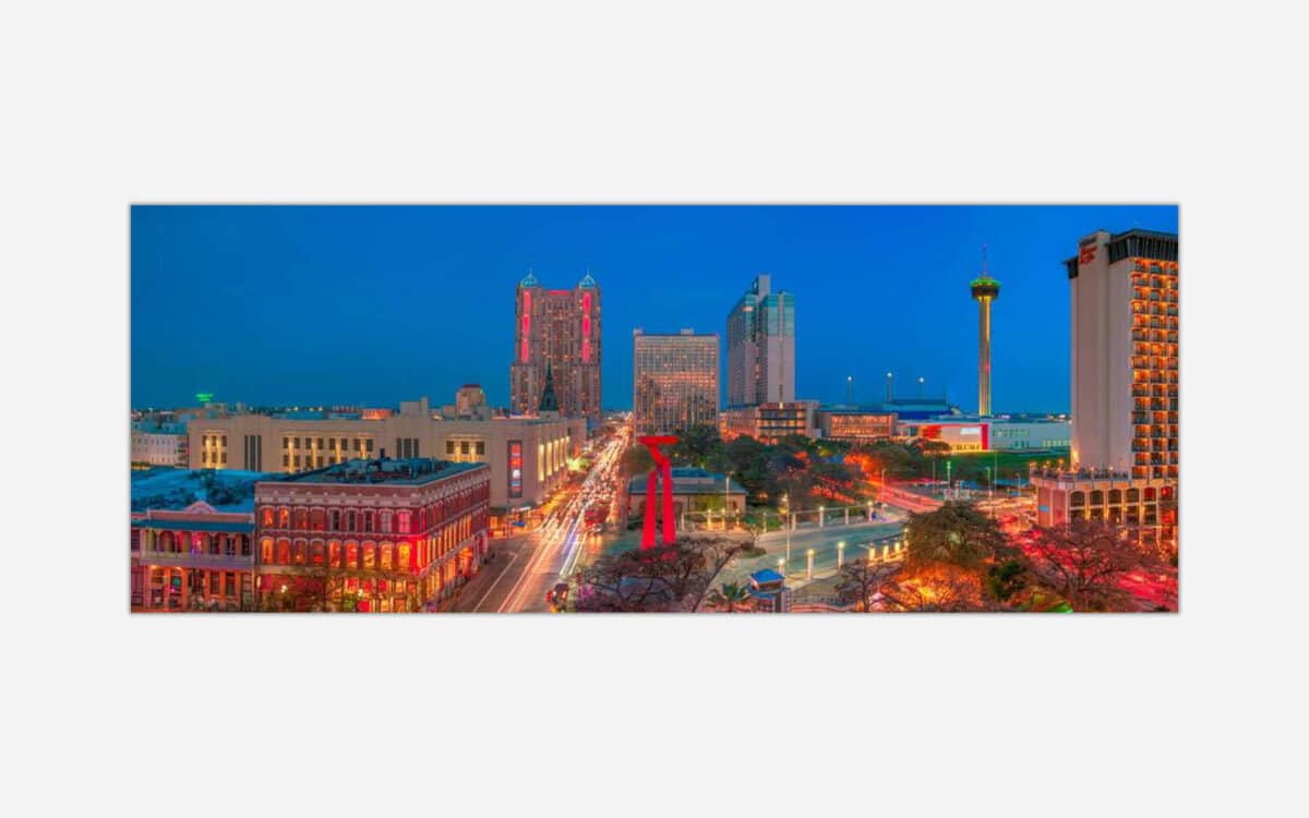 A panoramic view of an urban skyline at twilight with illuminated buildings, a tower, and traffic light trails.