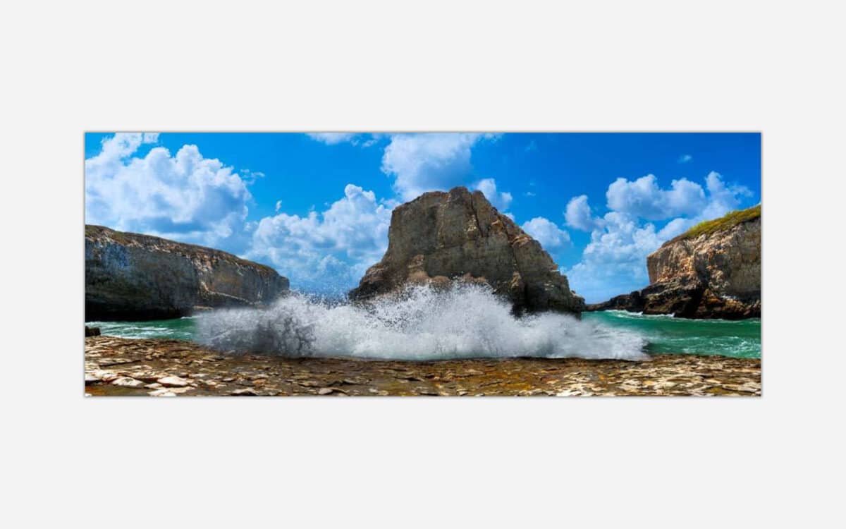 A panoramic photo of ocean waves crashing against rocky sea cliffs under a blue sky with fluffy clouds.