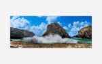 A panoramic photo of ocean waves crashing against rocky sea cliffs under a blue sky with fluffy clouds.