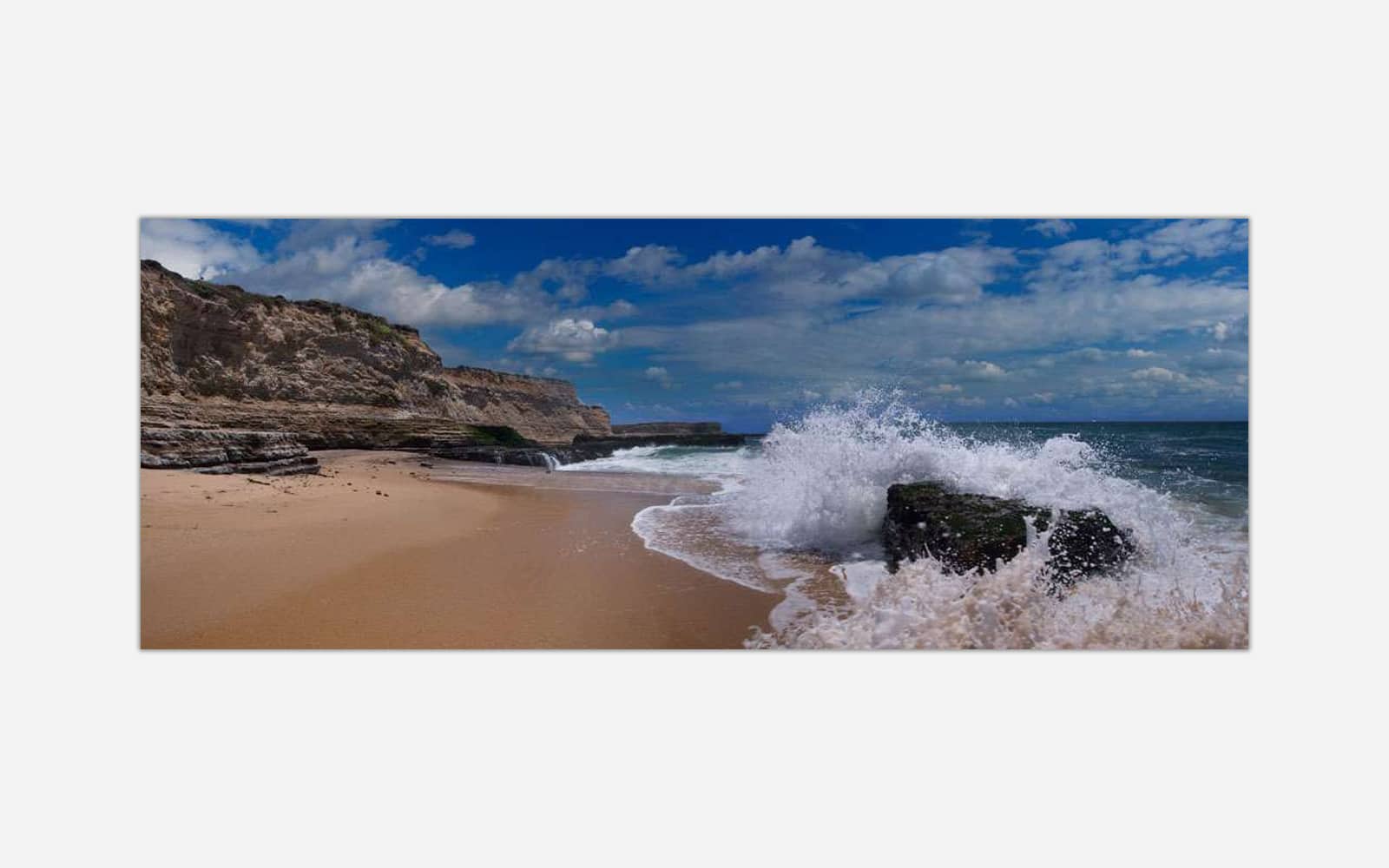 Splashed (1) Image shows a dynamic beach scene with a wave crashing against a rock on the sandy shore with a clear blue sky and coastal cliffs in the background.