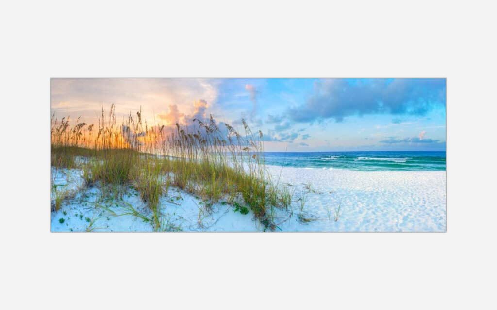 A panoramic wall art piece featuring a serene beach at sunset with ocean waves, fluffy clouds, and beach grass in the sand dunes.