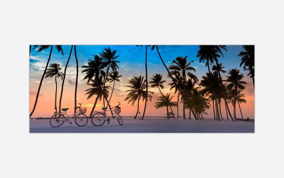 An art print of a serene beach sunset with silhouettes of palm trees and bicycles on white sand, under a pastel-colored sky.