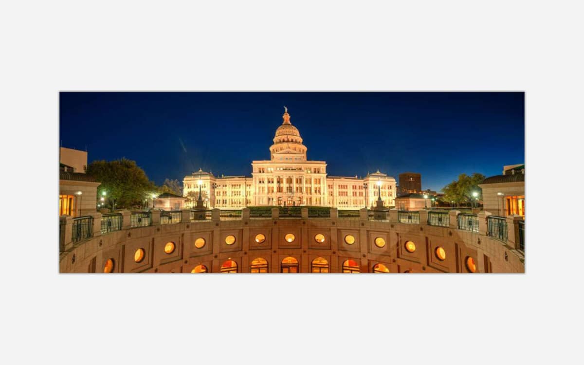 A nighttime view of a grand state capitol building with illuminated windows, surrounded by a cityscape under a twilight sky.