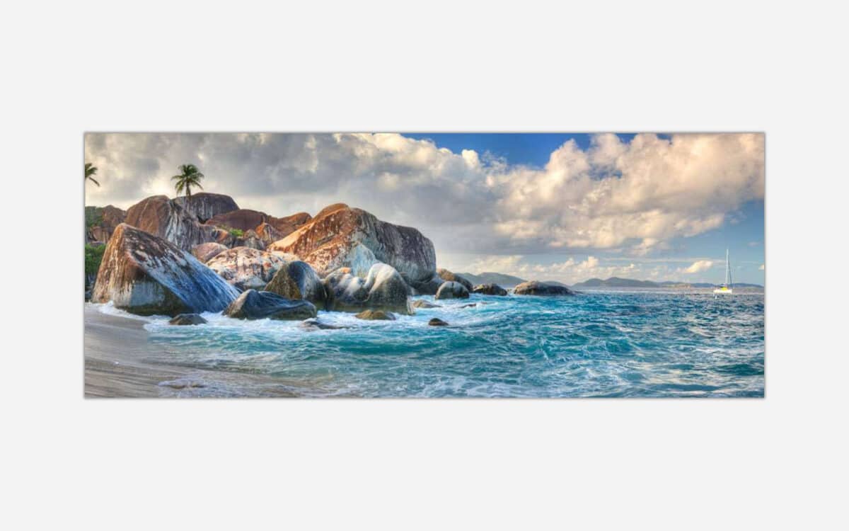 An image showing a sunny tropical beachscape with large rock formations, palm trees, blue ocean waters, and a distant sailboat under a partly cloudy sky.
