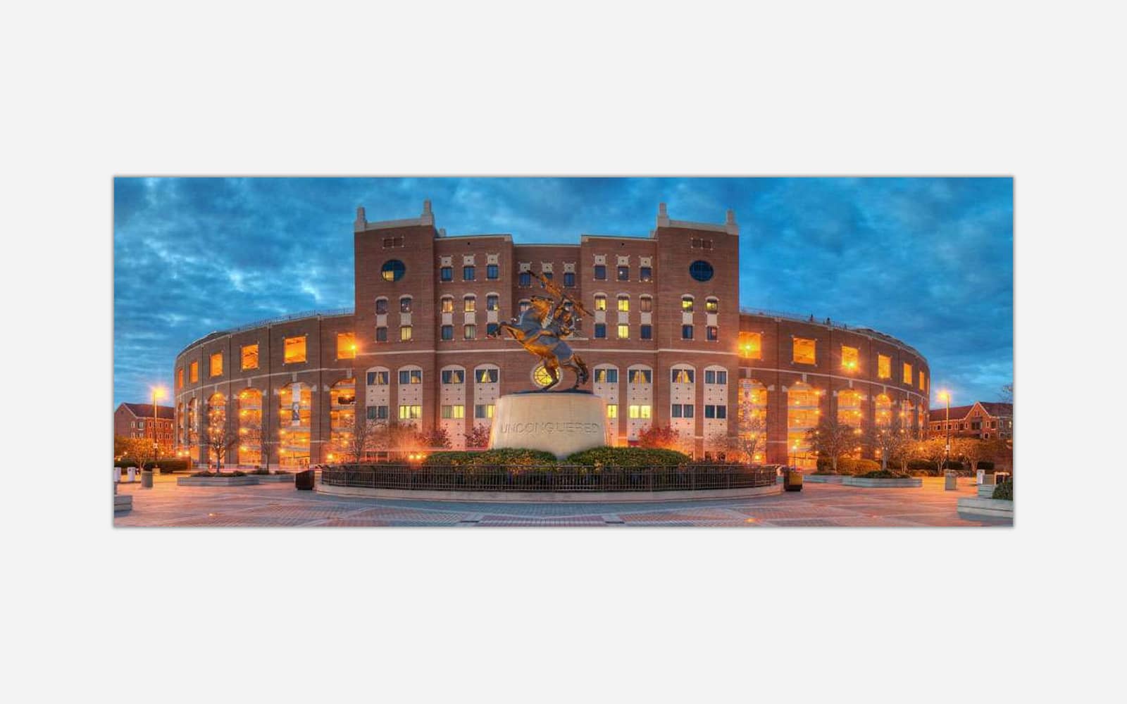 Unconquered (1) An image of a stadium taken at dusk with lights on, featuring a statue in front of the building and a blue sky in the background.