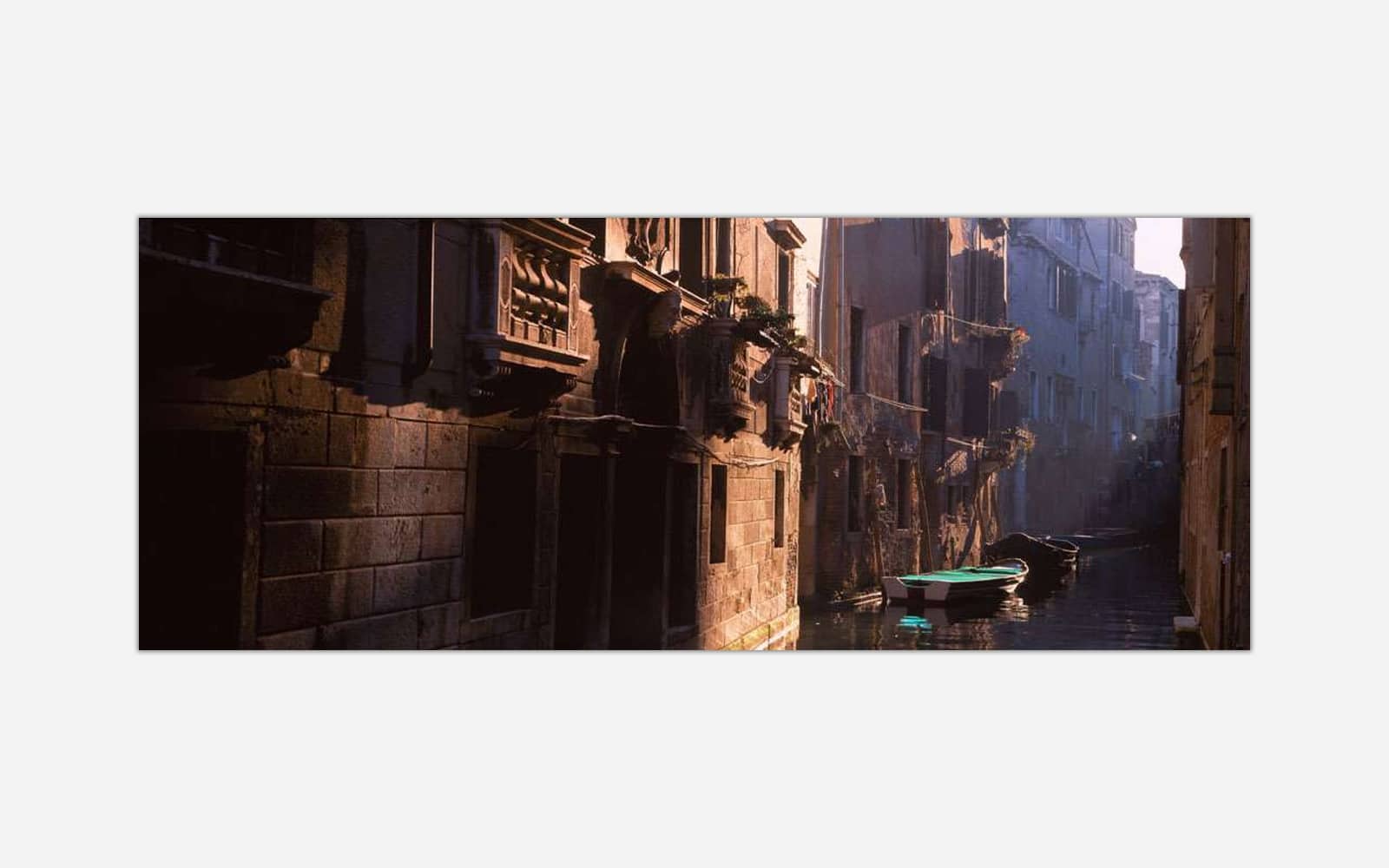 Venice (1) An image of a narrow Venice canal with historical buildings and a single boat, illuminated by soft sunlight.